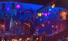 The image shows a vibrant theater set with colorful, illuminated lanterns, a bustling crowd of spectators, and a richly detailed stage scene reminiscent of a lively street, conveying the anticipation of a theatrical performance.