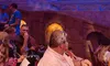 Audience members are seated in a theater with a vibrant and elaborate stage set resembling an exotic palace under blue lighting.
