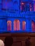 The image shows an illuminated stage set with gothic architecture elements, stained glass windows, and an audience in the foreground.