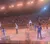 A group of performers in cowboy-themed costumes is entertaining an audience in an arena with a sandy floor.