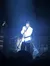 A performer in a white outfit illuminated by blue stage lighting appears to sing into a microphone with an audience in the foreground.