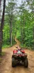 A red all-terrain vehicle is parked at the start of a winding dirt trail surrounded by a dense green forest.