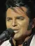 A person resembling Elvis Presley is singing into a microphone on a stage with a slot machine prop in the background.