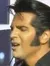 A performer emulating Elvis Presley sings into a microphone with a double bass player in the background on a stage lit with colorful lights.