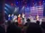 Performers are on stage singing in front of a large American flag backdrop with an audience in the foreground.