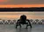 A solitary bell stands against a backdrop of a fiery sunset sky and tranquil water, framed by an ornate railing.