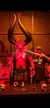 A person poses beside a large demonic figure with towering horns as part of a macabre and dramatic display, complete with skulls on a table in the foreground.