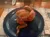 A roasted half chicken is presented on a dark blue plate, ready to be eaten at a restaurant with a reflective wooden table and a paper napkin in the background.