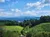 The image shows a lush green landscape with rolling hills in the foreground, a small town nestled in the valley, and layers of blue mountains in the distance under a partly cloudy sky.