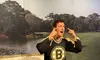 A life-sized figure is animatedly gesturing to its ears with a surprised expression, dressed in a Boston Bruins hockey jersey, against a backdrop of a tranquil golf course.