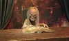 A creepy figure with a skull-like face and long hair is sitting behind a wooden table and appears to be reading a large, open book.