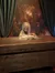 A creepy figure with a skull-like face and long hair is sitting behind a wooden table and appears to be reading a large, open book.