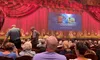 Audience members are seated in a theater with an ornate red curtain on stage and a screen displaying an advertisement for DVDs available in the lobby.