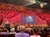 Audience members are seated in a theater with an ornate red curtain on stage and a screen displaying an advertisement for DVDs available in the lobby.