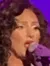 A person is performing on stage, singing into a microphone, wearing a sparkling dress with a purple-tinged spotlight shining on them.