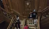 Visitors are ascending and descending a grand wooden staircase with ornate decorations in a lavish interior setting.