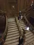 The image shows people descending a grand wooden staircase with ornate metal balustrades and a statue holding a lamp, while a person in a uniform stands at the bottom.
