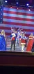 Performers in patriotic attire are singing and entertaining on stage, set against a large American flag backdrop.