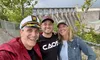 Three smiling individuals pose for a selfie with a large dam structure in the background.