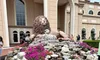 The image features a large, stately lion sculpture resting on a rock formation adorned with bright pink flowers, in front of an elegant building with archways, while visitors walk by and observe.