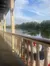 The image shows a river view from a balcony with decorative balusters, framed by lush greenery and a clear sky.