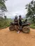 Two people wearing helmets are sitting on an all-terrain vehicle on a dirt track with a forested, mountainous backdrop.