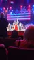 The image depicts a vibrant stage performance with performers dressed in patriotic American-themed costumes, singing or speaking before an audience, with a large American flag as a backdrop.