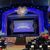The image shows the interior of a theater with a stage set for a performance titled