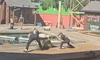 There are performers engaged in a lumberjack show, with one chopping wood and another taking aim with a crosscut saw, in a rustic outdoor setting implying a competitive atmosphere.