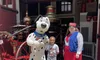 A child stands smiling between a person in a dalmatian costume and another person dressed in a red apron and striped shirt, in front of an antique fire engine at a themed park or event.