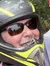 Two people wearing motorcycle helmets are smiling for a selfie outdoors, with one wearing a red helmet and the other a yellow and black one.
