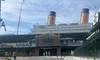 The image shows a sunny day at an exhibition or themed attraction modeled after the Titanic, with people strolling near the entrance.
