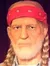The image shows a life-sized figure, resembling an older man with braided hair and wearing a red cap, a plaid shirt, and jeans, staged in front of a red curtain alongside an informational display and a musical instrument, likely a waxwork or a statue, designed to mimic a person's appearance.