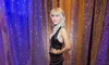 A figure, likely a wax figure, resembling a woman with blonde hair in a black dress, poses confidently in front of glittering multicolored curtains with sparkling light reflections on the floor.