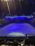 The image shows an indoor arena set up for an event with an ice floor, artificial rocky terrains, theatrical lighting, and an audience seated around the stage area, with the text 
