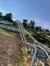 The image shows a metal alpine coaster track winding down a hillside, surrounded by greenery, with a blue sky above and a white fence running parallel to the coaster track at the base of the hill.