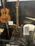 The image shows a unique, ornately carved electric guitar on display, surrounded by other vintage musical instruments inside a glass case.