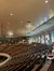 The photo shows the interior of a spacious auditorium or theater with rows of wooden seats, colorful stained glass windows, and a high wooden ceiling with mounted lights.