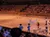 The image shows a performance in a sandy arena with people in western-style costumes, possibly part of a rodeo or themed show, being watched by a large audience seated in tiered rows.