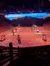The image shows a group of performers in a sand-filled arena with a rustic backdrop, entertaining an audience in a dimly-lit amphitheater.