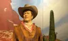 A wax figure of a cowboy, complete with a hat, bandana, and rifle, stands against a backdrop of a desert scene with a cactus.