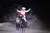 A person in an ornate costume is riding a horse and gesturing grandly with their arms outstretched in an illuminated arena setting.
