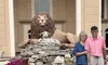 In front of an elegant building, a smiling pair stands by a large sculpture of a lion and a lamb resting on a rocky base adorned with flowers.