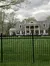 The image shows a large, stately two-story home with a white facade, multiple windows, and a central portico, surrounded by trees and secured behind a black metal fence, under a cloudy sky.