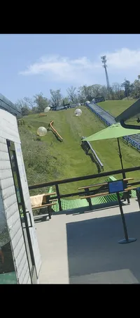 The image shows an outdoor activity area with large inflatable transparent balls that people can go inside of, set on a grassy hill for rolling down, with a sign that reads