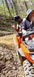 Two individuals are riding an orange ATV on a dirt trail with leafy trees in the background, and the passenger gestures while wearing a motorcycle helmet.