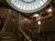 The image shows an opulent wooden staircase with ornate metal railings, an angel statue holding a lamp, and a circular skylight, conveying a sense of vintage luxury.