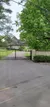 The image shows a view of a gated driveway leading to a large, two-story house, surrounded by green trees and a landscaped lawn, with power lines overhead.