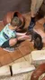 A child is crouched on the floor offering food to a small rodent-like animal inside an indoor setting with terracotta pots and onlookers.