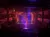 The image depicts a live performance on a stage with vibrant lighting, watched by an audience in a multi-tiered theater.